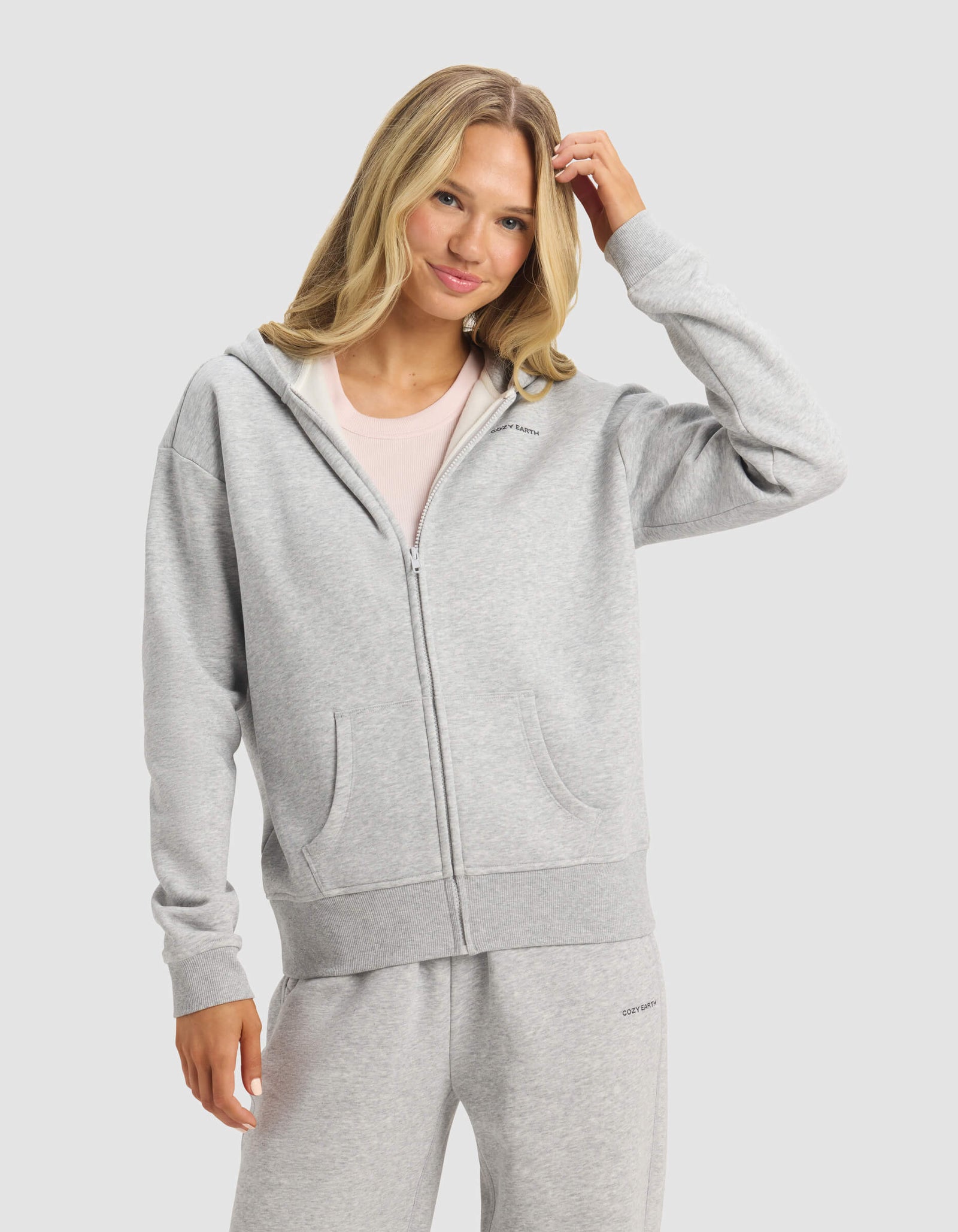 A woman with long blonde hair smiles and touches her hair while wearing Cozy Earth's Women's CityScape 2.0 Full Zip hoodie and matching sweatpants, standing against a plain light background.