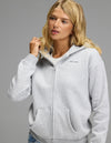 A woman with long blonde hair poses against a plain gray background, wearing the Cozy Earth Women’s CityScape Full Zip Hoodie in Heather Grey over a white shirt, with one hand resting near the collar.