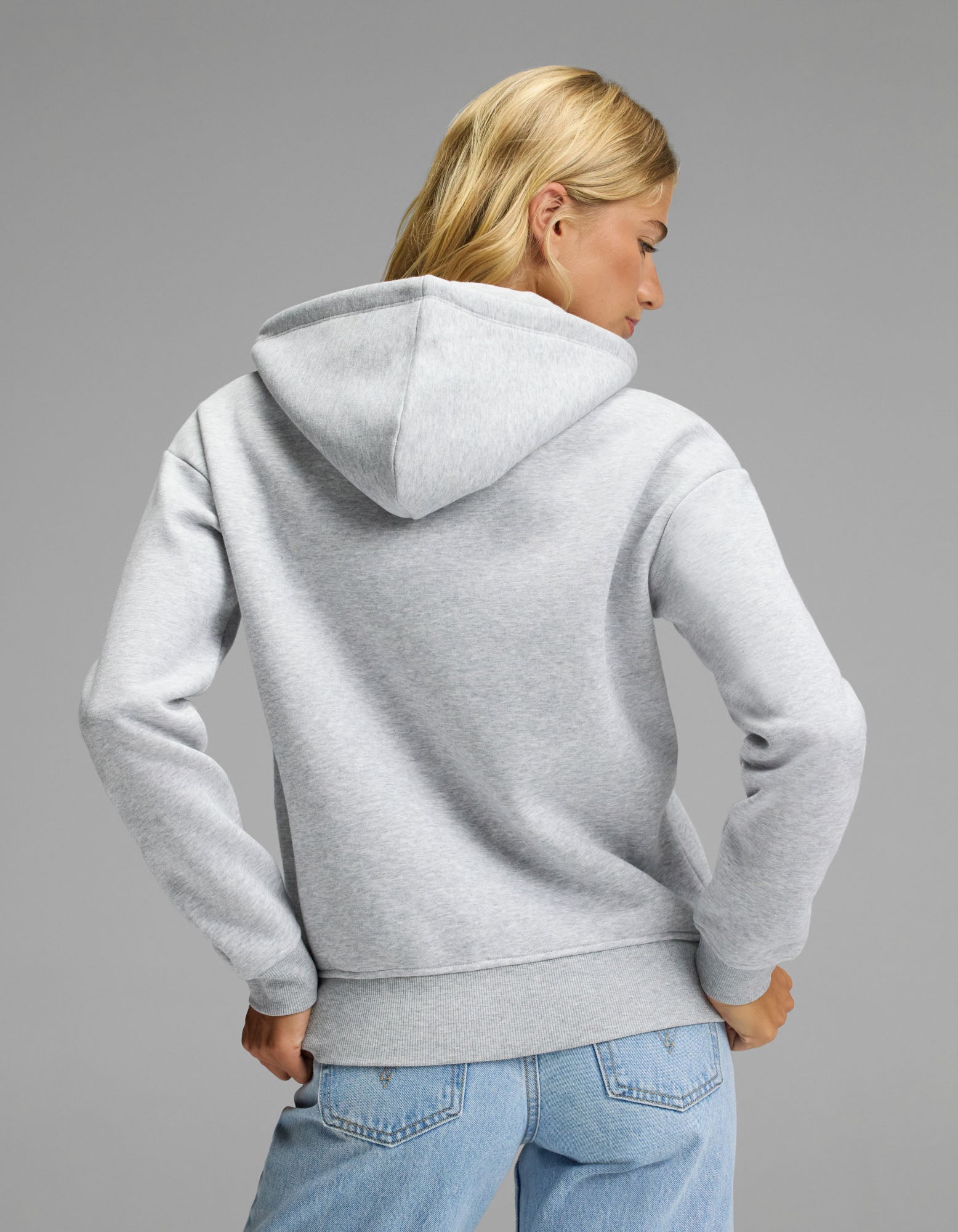 A woman with blonde hair, wearing the Cozy Earth Women’s CityScape 2.0 Full Zip Hoodie in light gray and blue jeans, stands facing away from the camera against a plain gray background.
