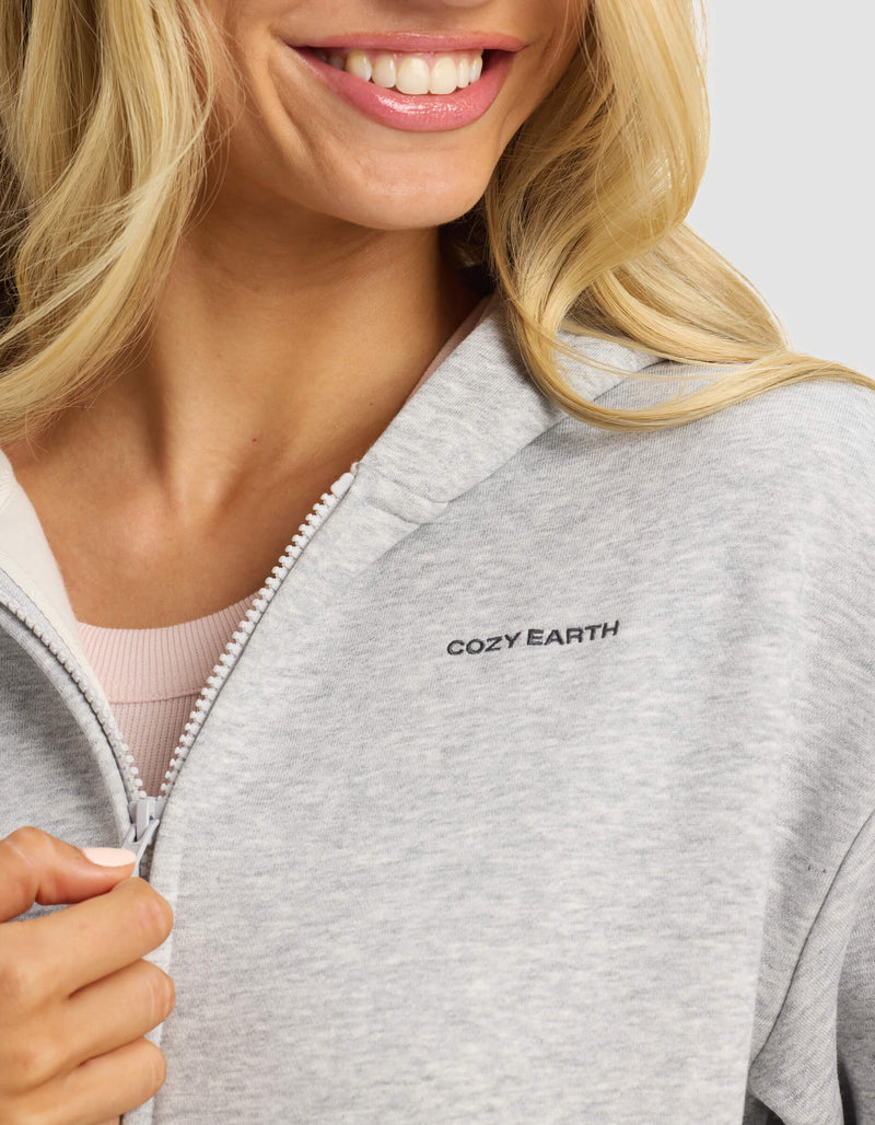 A woman with long blonde hair smiles, holding the zipper of a light gray Women's CityScape 2.0 Full Zip hoodie by Cozy Earth. Only the lower half of her face is shown, and "COZY EARTH" is printed on the chest.