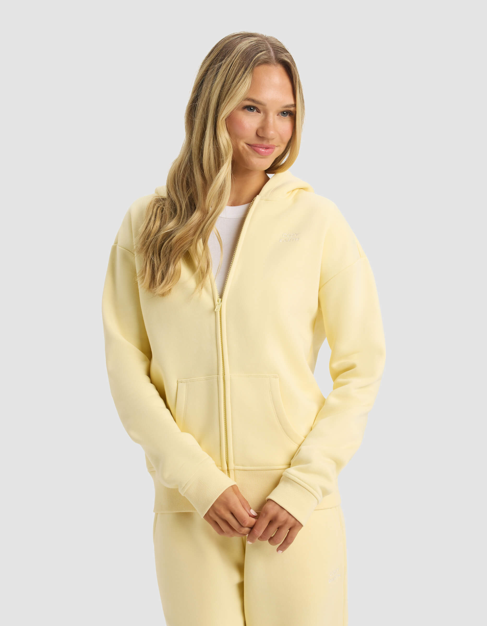 A woman with long blonde hair wears the Cozy Earth Women's CityScape 2.0 Full Zip hoodie and matching sweatpants, standing with her hands together and smiling slightly against a plain light gray background.