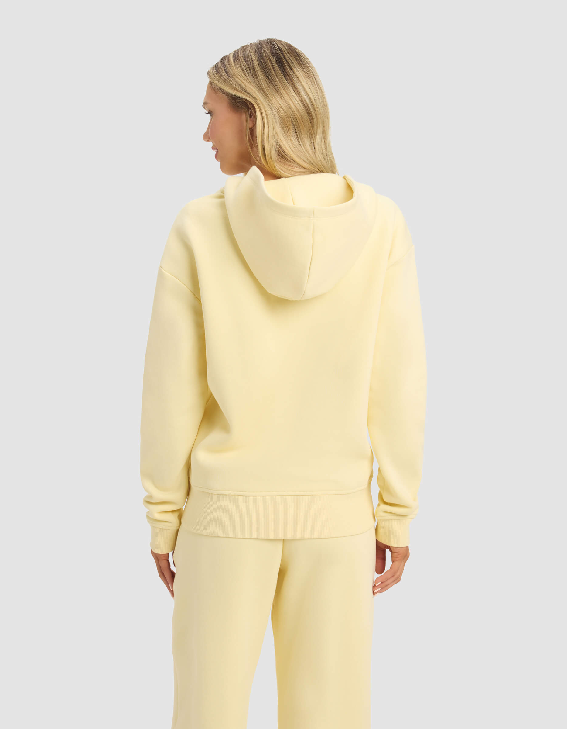 A person with long blonde hair is seen from behind wearing the Cozy Earth Women's CityScape 2.0 Full Zip hoodie and matching pants in pale yellow, standing against a plain light gray background.