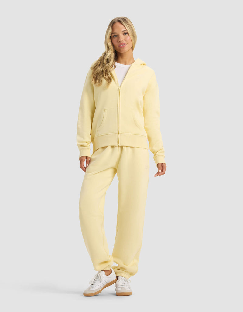 A woman with long blonde hair smiles softly against a gray background, wearing the Cozy Earth Women's CityScape 2.0 Full Zip hoodie in light yellow, matching sweatpants, a white shirt, and white sneakers.