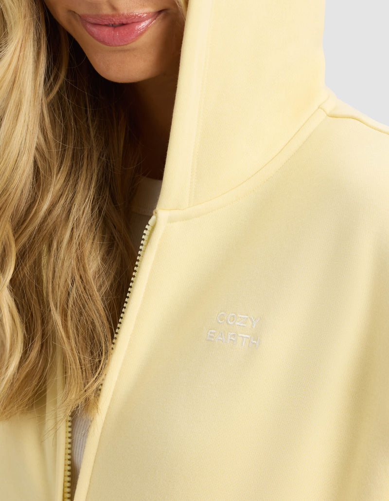 A close-up shows a woman wearing the Cozy Earth Women's CityScape 2.0 Full Zip hoodie in light yellow, with the hood up and only her lower face and blond hair visible. "COZY EARTH" is embroidered in white on the chest.