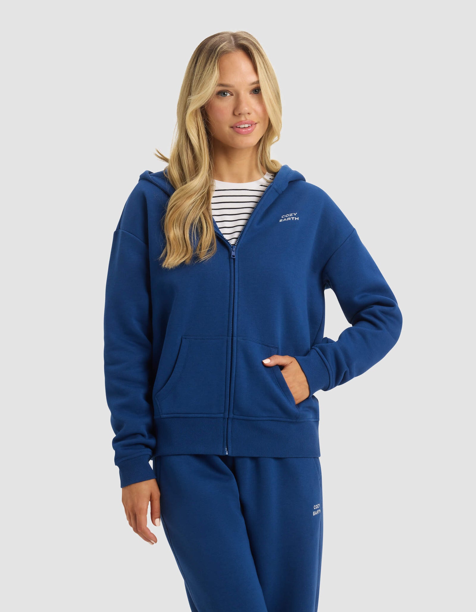 A woman with long blonde hair models the Cozy Earth Women's CityScape 2.0 Full Zip hoodie and matching blue sweatpants over a white and navy striped shirt, posing with one hand in her pocket against a plain light background.