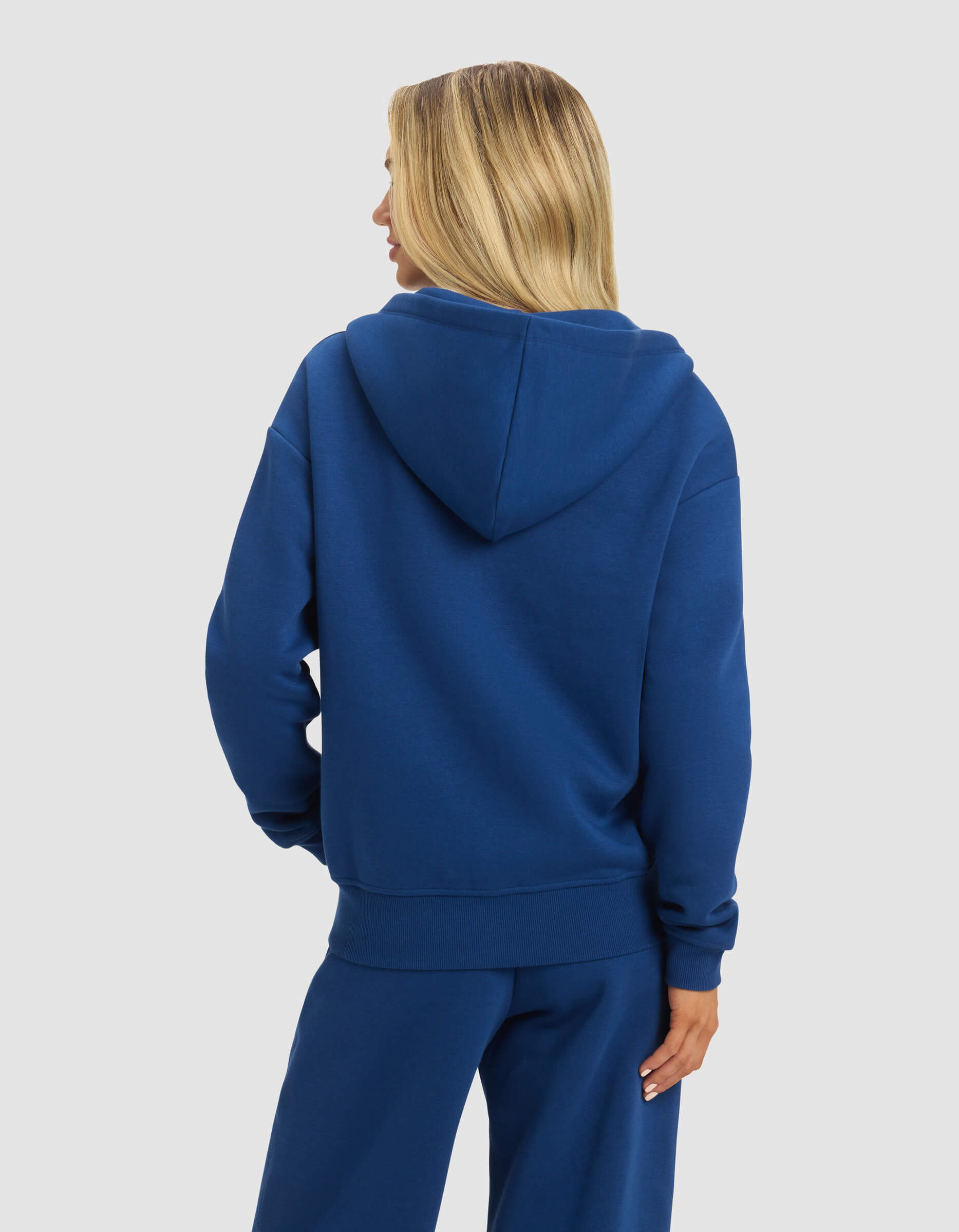 A person with long blonde hair is shown from the back, wearing the Cozy Earth Women's CityScape 2.0 Full Zip in blue with matching pants against a plain light gray background.