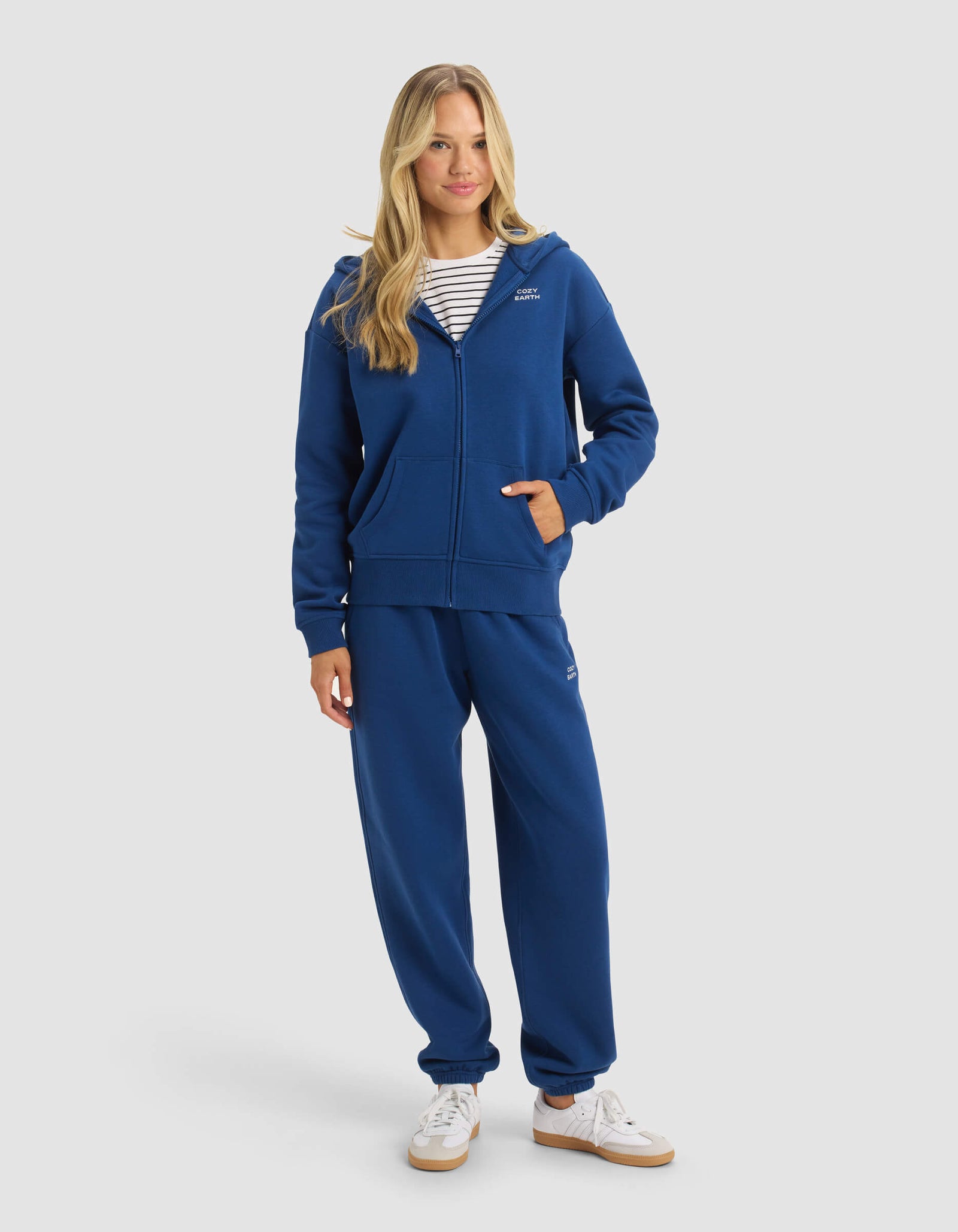 A woman with long blonde hair smiles slightly against a plain background, wearing Cozy Earth's Women's CityScape 2.0 Full Zip hoodie in blue, matching sweatpants, a striped shirt underneath, and white sneakers.