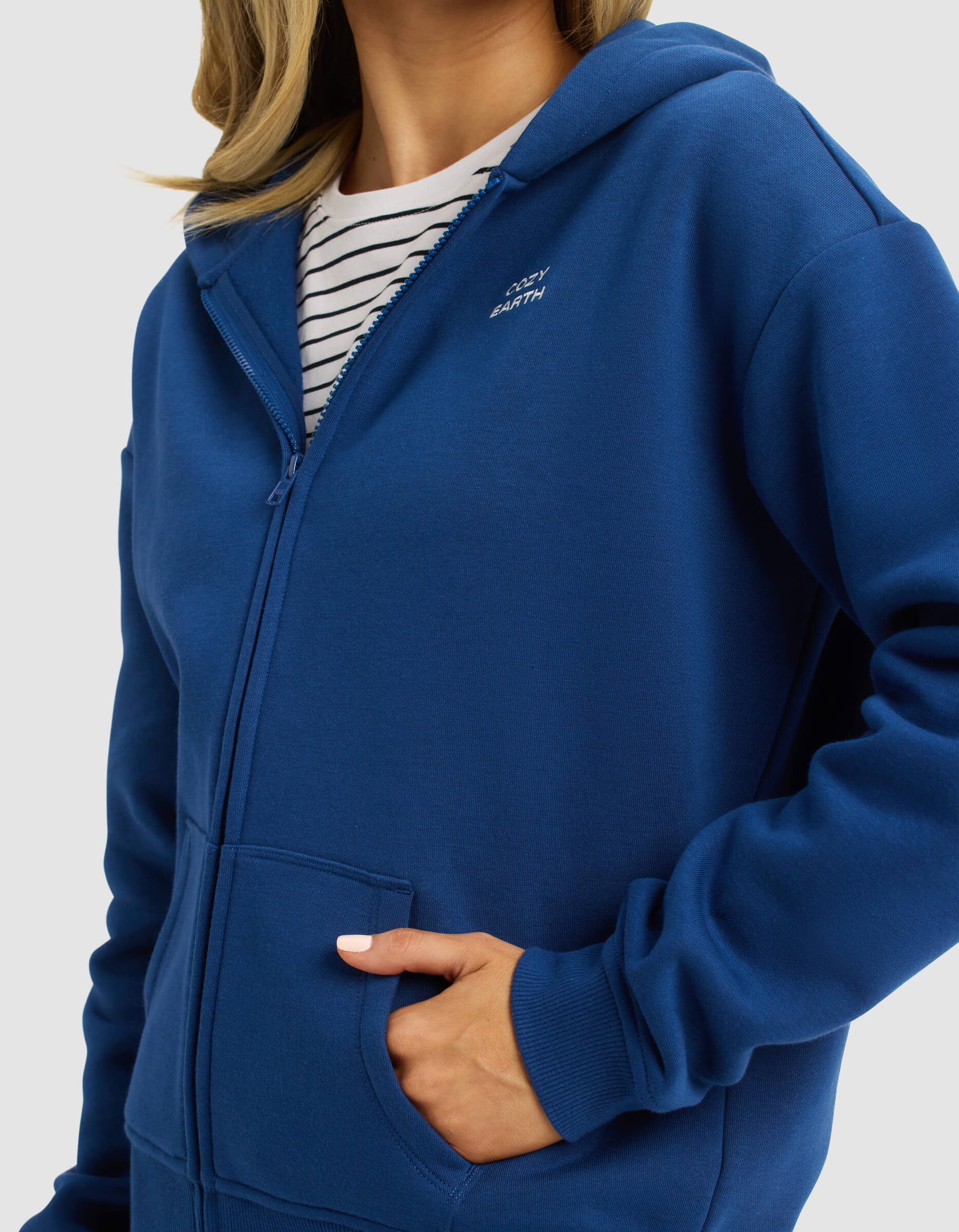A woman wears the Cozy Earth Women's CityScape 2.0 Full Zip hoodie in blue over a white and black striped shirt, with one hand in the pocket, shown from shoulders to hips against a plain background.