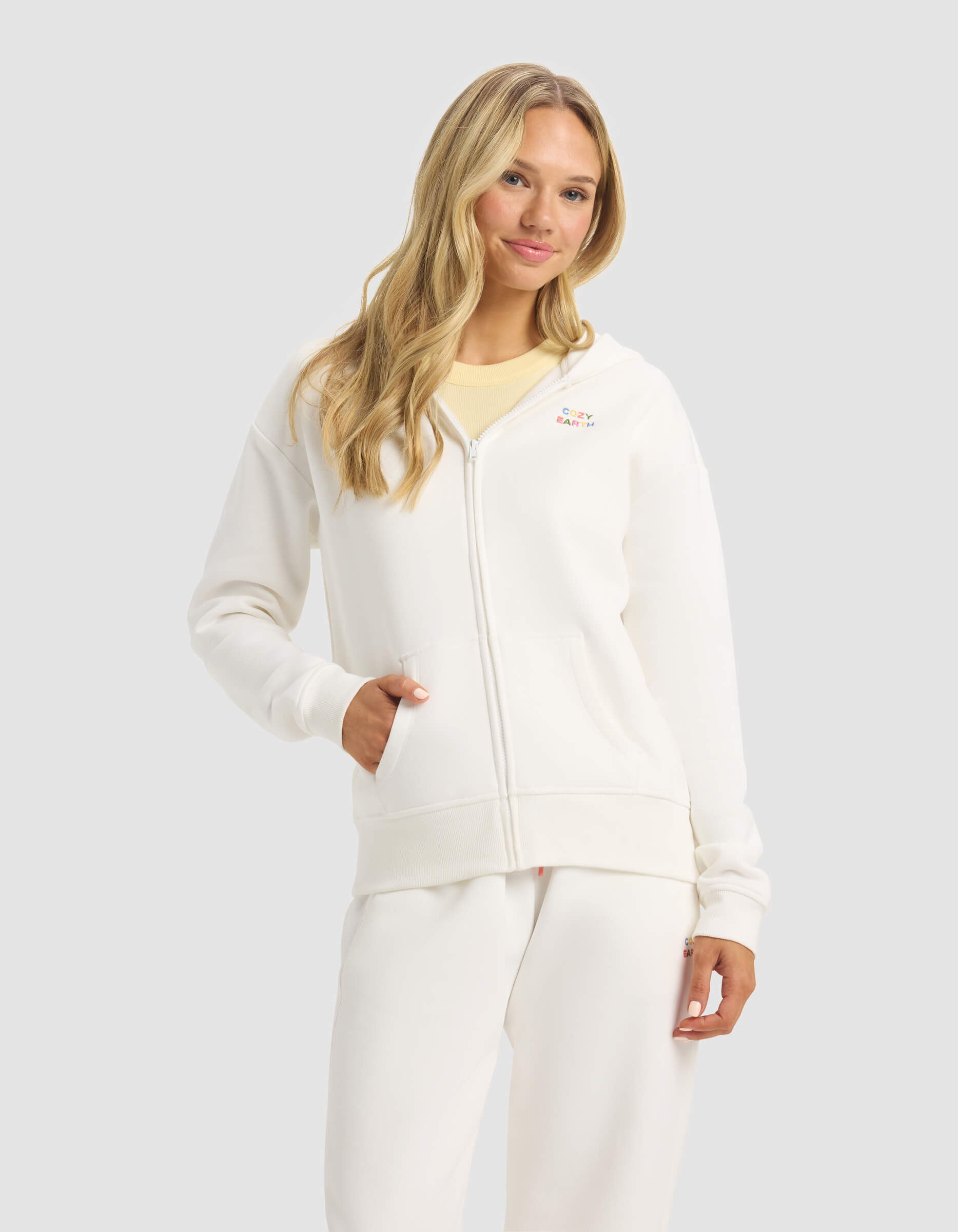 A woman with long blonde hair models the Cozy Earth Women's CityScape 2.0 Full Zip hoodie and matching white sweatpants against a light gray background, one hand in her hoodie pocket.