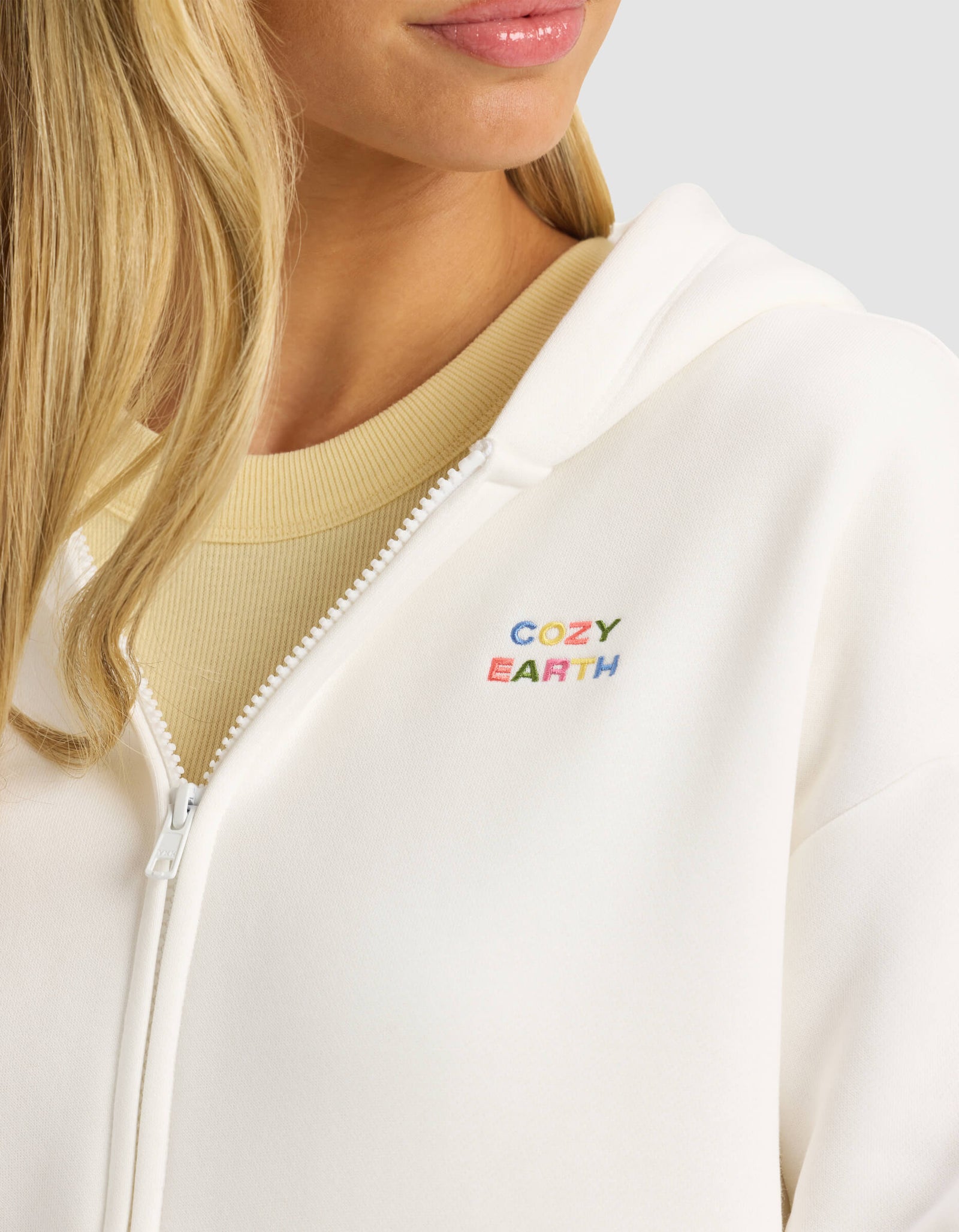A woman wears the Cozy Earth Women's CityScape 2.0 Full Zip—a white hoodie with colorful "COZY EARTH" embroidery on the chest—over a beige top. Only her lower face, neck, and part of her blonde hair are visible.
