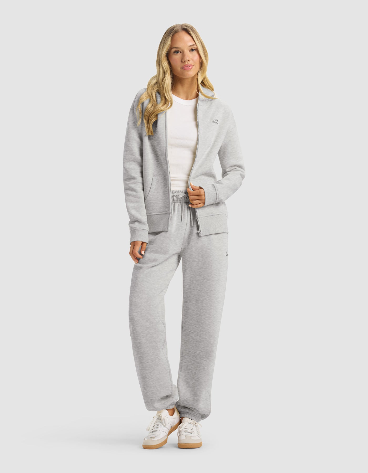 A woman with long blonde hair wears the Cozy Earth Women's CityScape 2.0 Full Zip Hoodie & Sweatpant Set in light gray, paired with a white t-shirt and sneakers, standing relaxed against a plain light background.