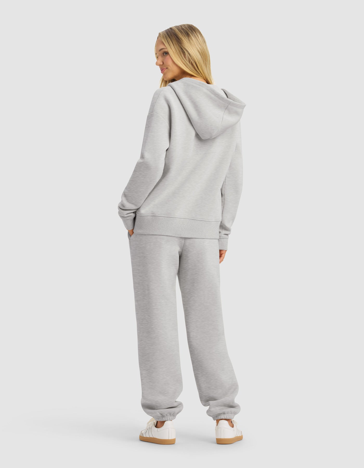 A woman with long blonde hair models the Cozy Earth Women's CityScape 2.0 Full Zip Hoodie & Sweatpant Set in light gray, facing away and looking over her shoulder on a plain white background, paired with white sneakers.