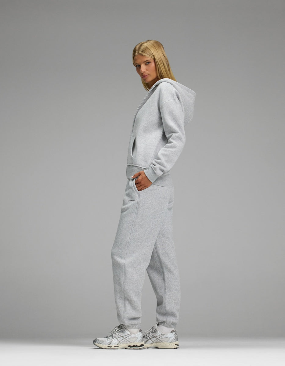 Model wears the Women’s CityScape Full-Zip Hoodie & Sweatpant Set by Cozy Earth in light gray with white sneakers, standing against a plain gray background.