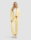 A woman with long blonde hair wears the Cozy Earth Women's CityScape 2.0 Full Zip & Sweatpant Set in light yellow, paired with a white t-shirt and sneakers. She stands facing forward against a plain light gray background.|Color:Meringue