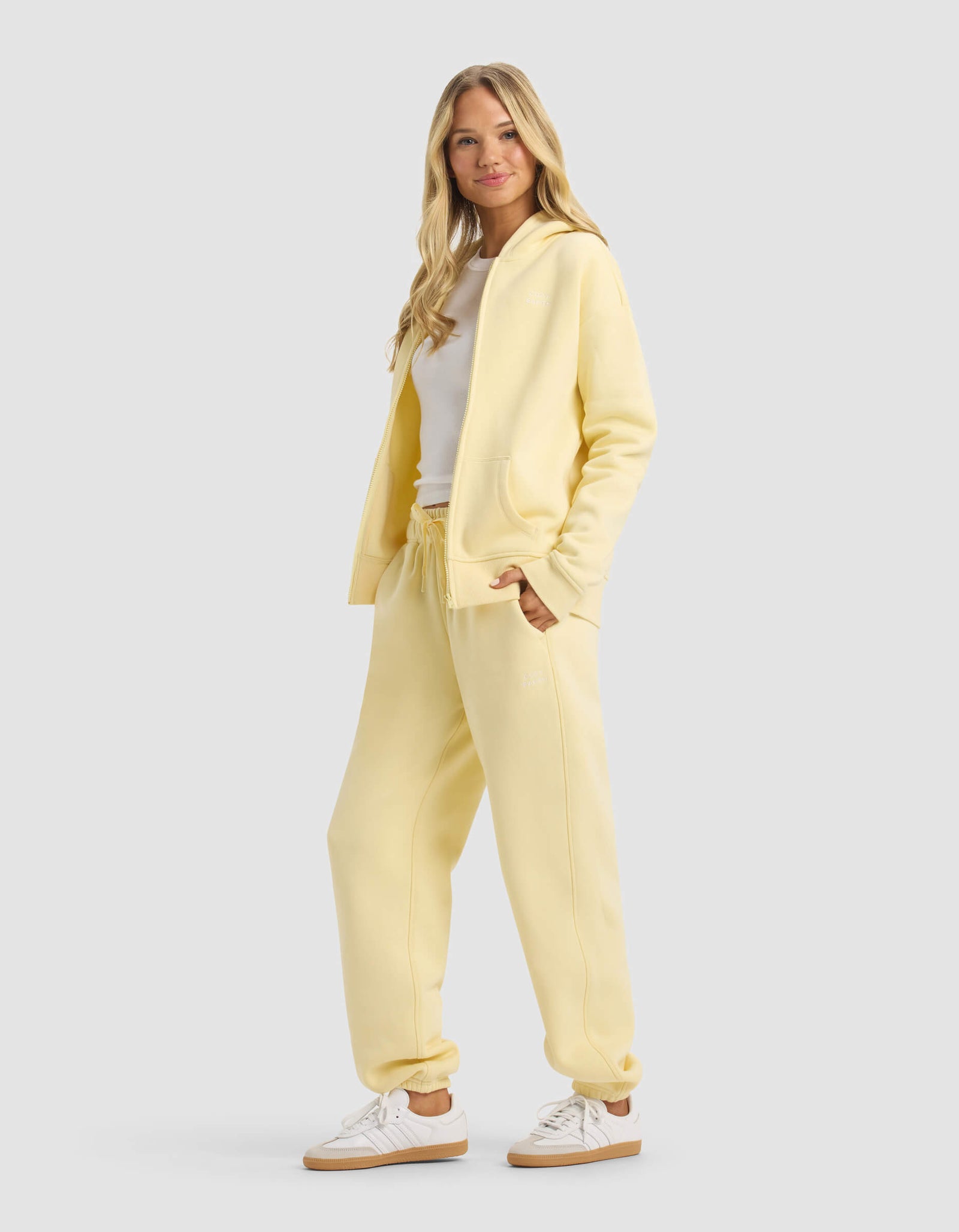 A woman with long blonde hair smiles in Cozy Earth’s Women's CityScape 2.0 Full Zip & Sweatpant Set, paired with white sneakers and a white top, standing against a plain light gray background.