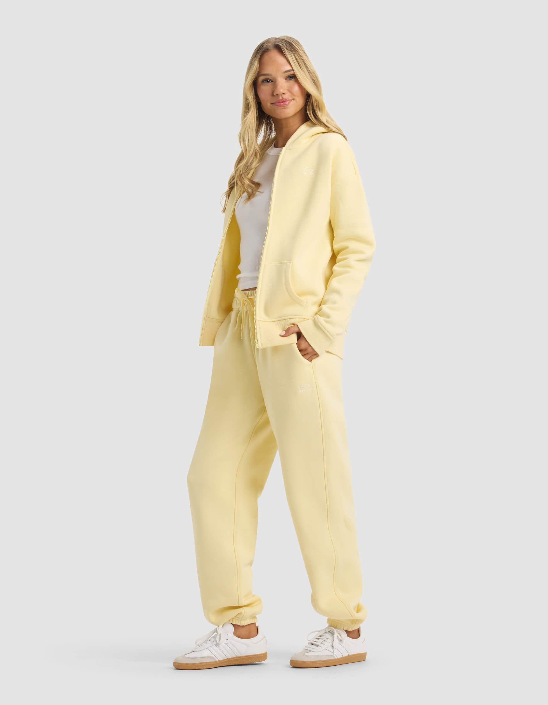 A woman with long blonde hair smiles in Cozy Earth’s Women's CityScape 2.0 Full Zip & Sweatpant Set, paired with white sneakers and a white top, standing against a plain light gray background.|Color:Meringue