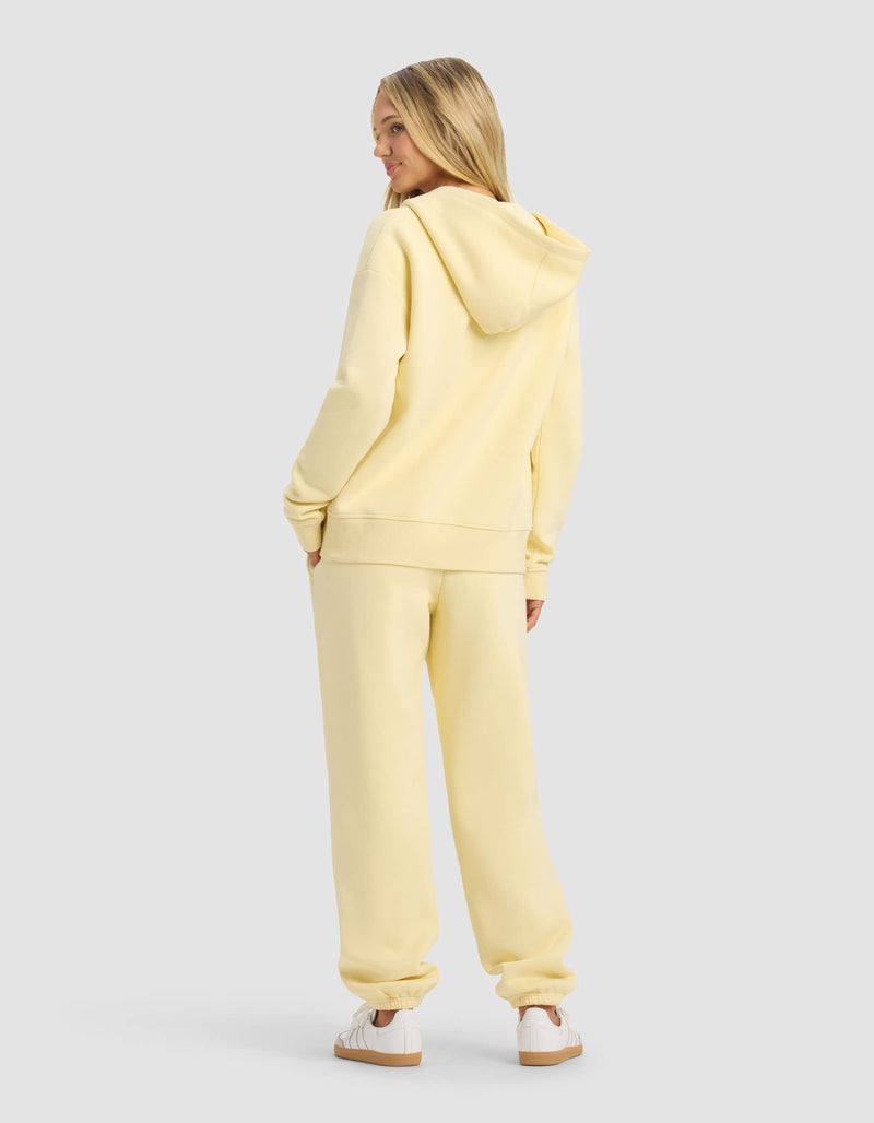 A woman with long blonde hair faces away, wearing the Cozy Earth Women's CityScape 2.0 Full Zip & Sweatpant Set in light yellow and white sneakers, posed against a plain light gray background.