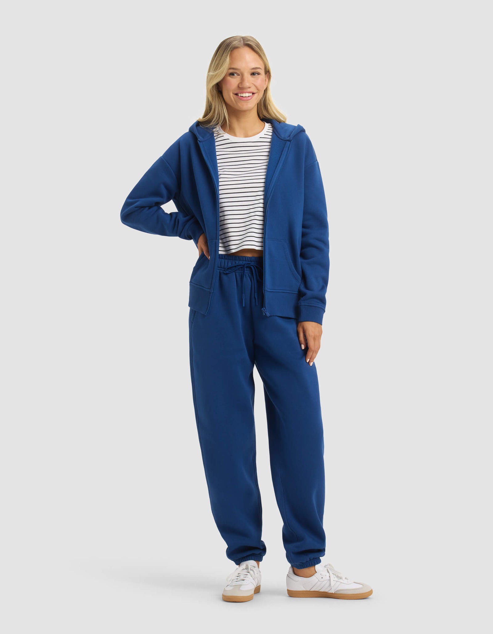 A woman smiles in Cozy Earth's Women's CityScape 2.0 Full Zip & Sweatpant Set—blue zip-up hoodie and matching sweatpants—paired with a white and navy striped tee and white sneakers, set against a light gray background.