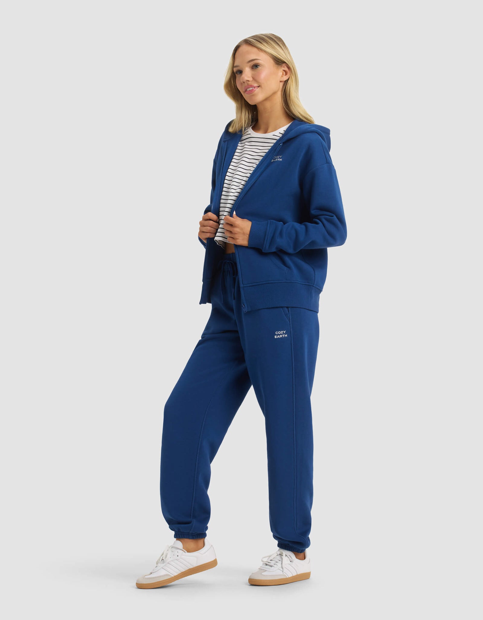 A woman wears the Cozy Earth Women's CityScape 2.0 Full Zip & Sweatpant Set, paired with white sneakers and a striped shirt, smiling and looking to the side against a plain light background.