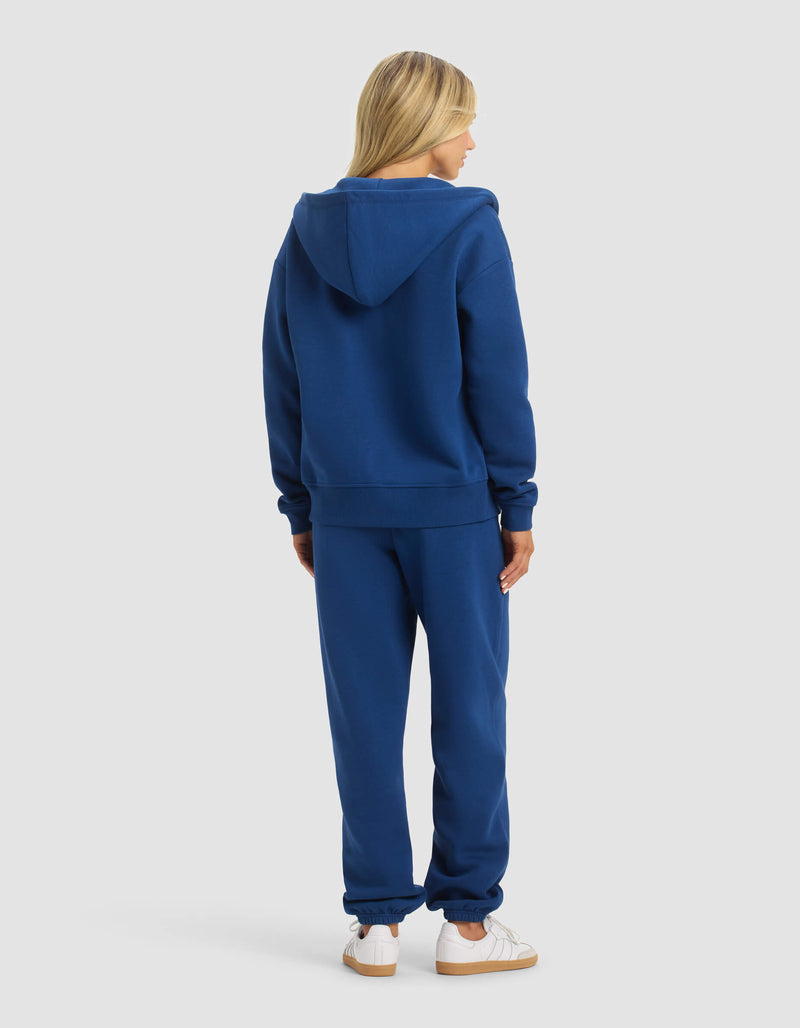 A woman with blonde hair stands facing away, wearing Cozy Earth's Women's CityScape 2.0 Full Zip & Sweatpant Set in royal blue with white sneakers, set against a plain light gray background.