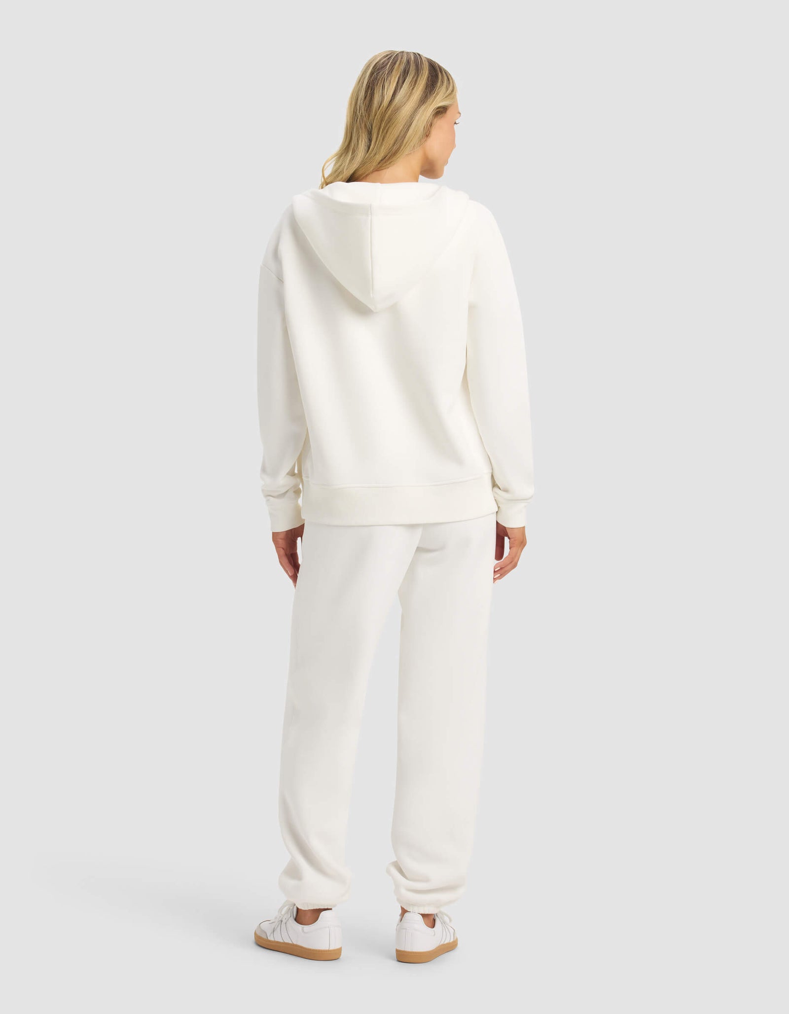 A woman with blonde hair faces away, wearing the Cozy Earth Women's CityScape 2.0 Full Zip & Sweatpant Set in white with matching sneakers, against a plain light gray background.