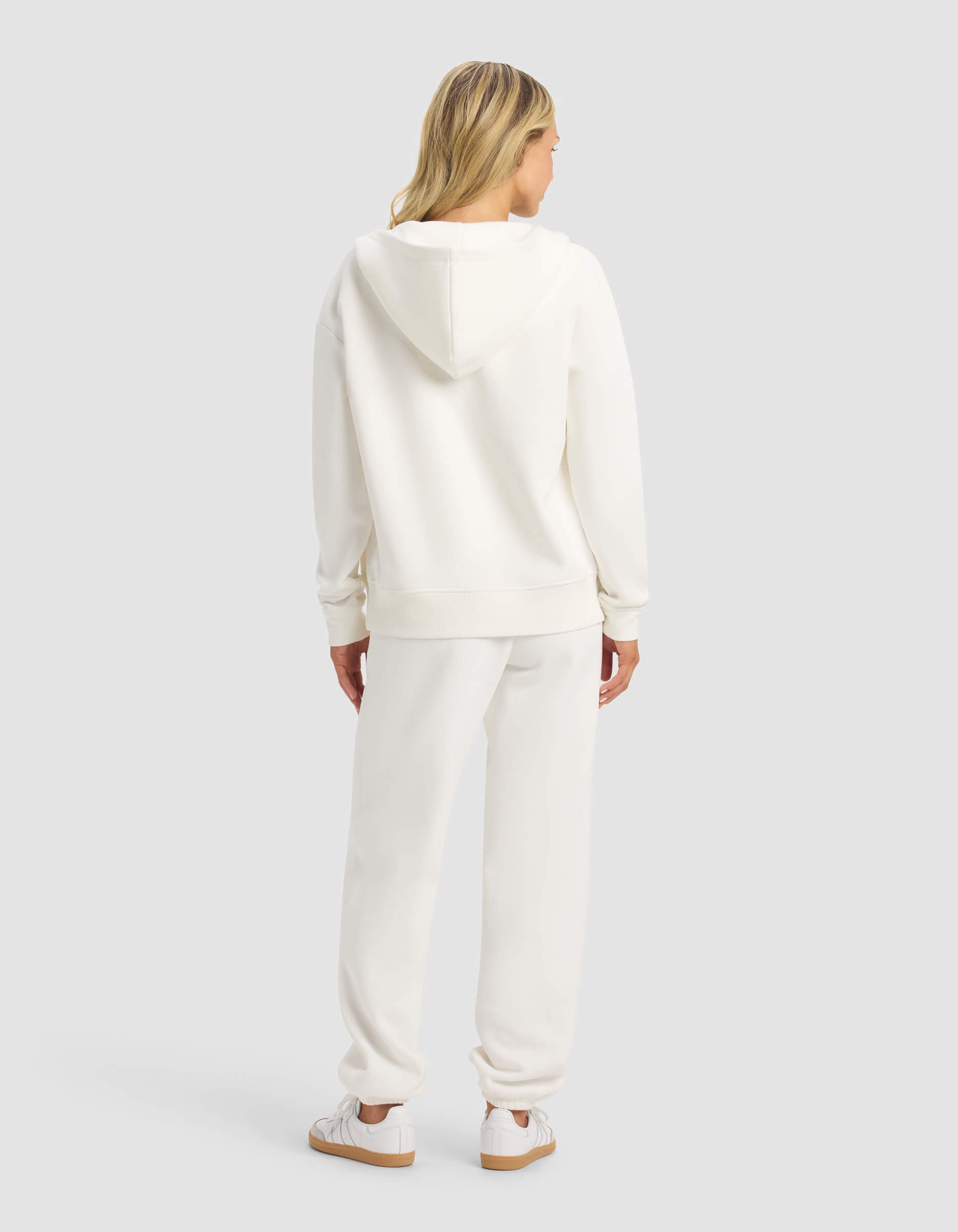 A woman with blonde hair faces away, wearing the Cozy Earth Women's CityScape 2.0 Full Zip & Sweatpant Set in white with matching sneakers, against a plain light gray background.|Color:Sea Salt