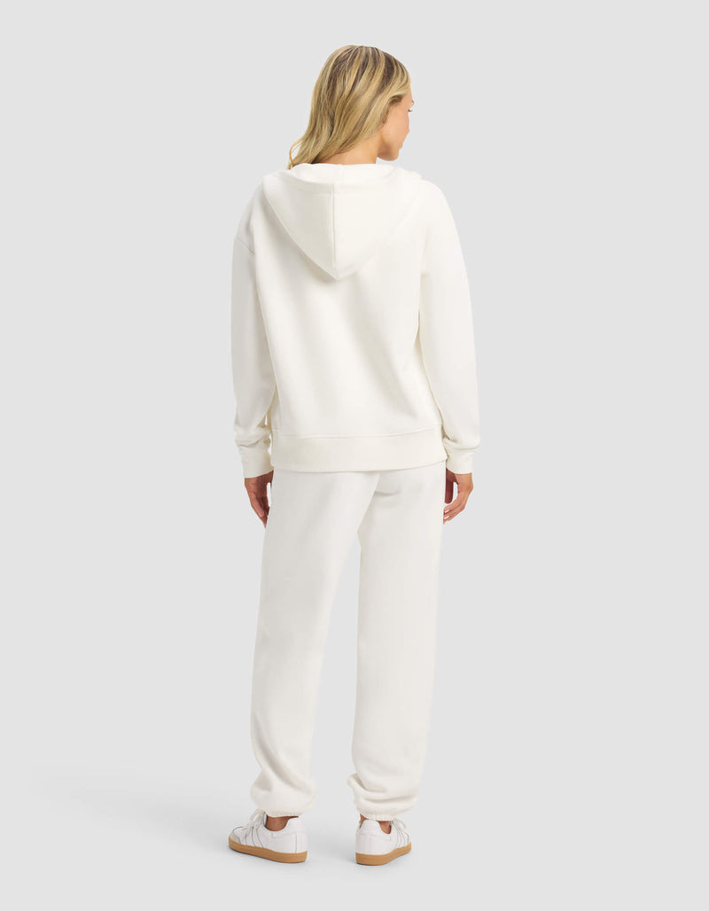 A woman with blonde hair faces away, wearing the Cozy Earth Women's CityScape 2.0 Full Zip & Sweatpant Set in white with matching sneakers, against a plain light gray background.