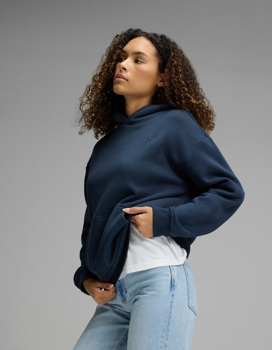 Wearing the Cozy Earth Women’s CityScape Hoodie in Eclipse over a white shirt and light blue jeans, a person with curly hair stands against a plain gray background, gazing slightly upward.