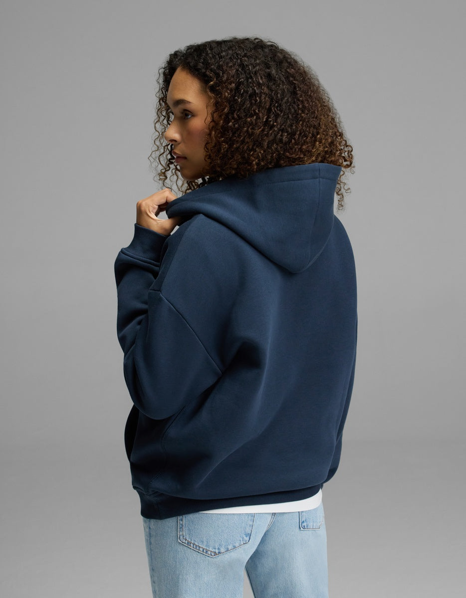 A woman with curly hair, dressed in the Cozy Earth Women’s CityScape Hoodie in navy blue and light blue jeans, stands facing away and gazes to the side against a plain gray background.|Color:Eclipse