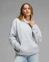 A woman models the Cozy Earth Women’s CityScape Hoodie in Heather Grey, pairing it with light blue jeans as she stands against a plain gray background—one hand adjusting the hoodie’s collar, the other tucked into its front pocket.|Color:Heather Grey