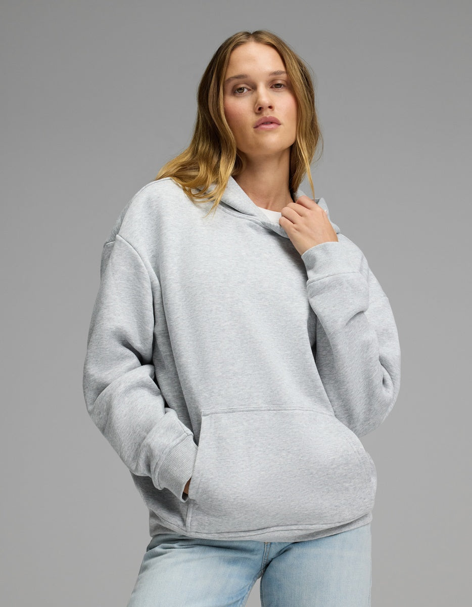 A woman models the Cozy Earth Women’s CityScape Hoodie in Heather Grey, pairing it with light blue jeans as she stands against a plain gray background—one hand adjusting the hoodie’s collar, the other tucked into its front pocket.