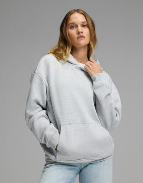 A woman models the Cozy Earth Women’s CityScape Hoodie in Heather Grey, pairing it with light blue jeans as she stands against a plain gray background—one hand adjusting the hoodie’s collar, the other tucked into its front pocket.|Color:Heather Grey