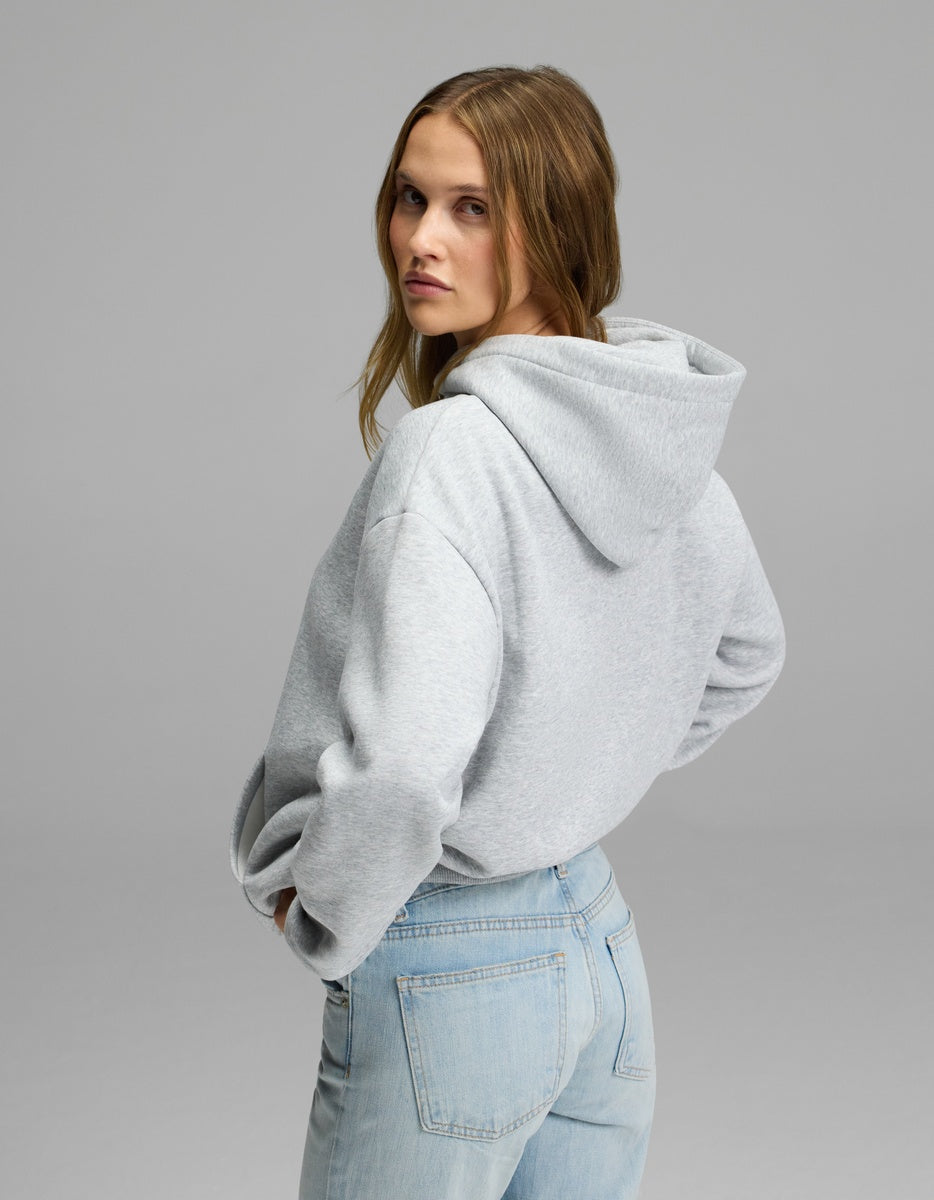 A woman with light brown hair models the Cozy Earth Women’s CityScape Hoodie in light gray, paired with light blue jeans. She stands turned slightly to the side, glancing back over her shoulder against a plain gray background.