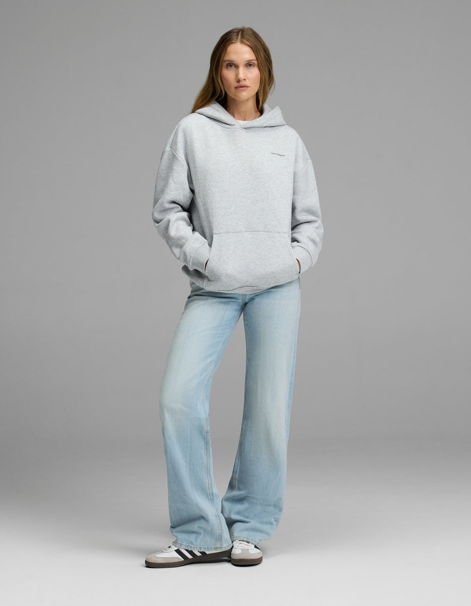 A woman stands against a plain gray background, wearing the Cozy Earth Women’s CityScape Hoodie, paired with light blue jeans and white sneakers with black stripes. Her hands rest in the hoodie’s pocket as she looks straight ahead with a neutral expression.