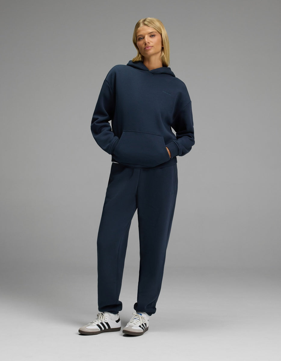 A woman stands in a relaxed pose wearing the Cozy Earth Women’s CityScape Hoodie & Sweatpant Set in navy blue, paired with white sneakers against a plain gray background.