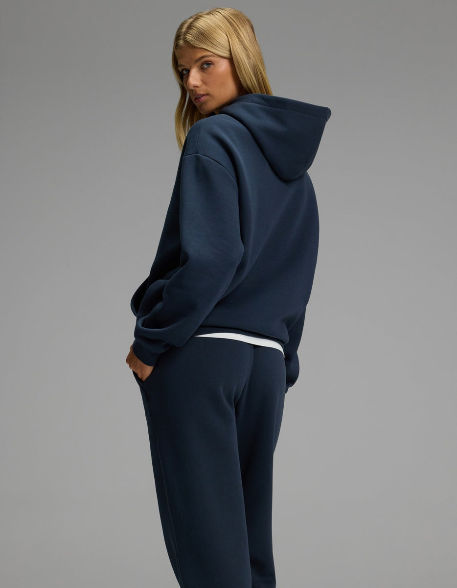 A woman with long blonde hair models the Cozy Earth Women’s CityScape Hoodie & Sweatpant Set in navy blue, standing sideways and looking over her shoulder against a plain gray background.