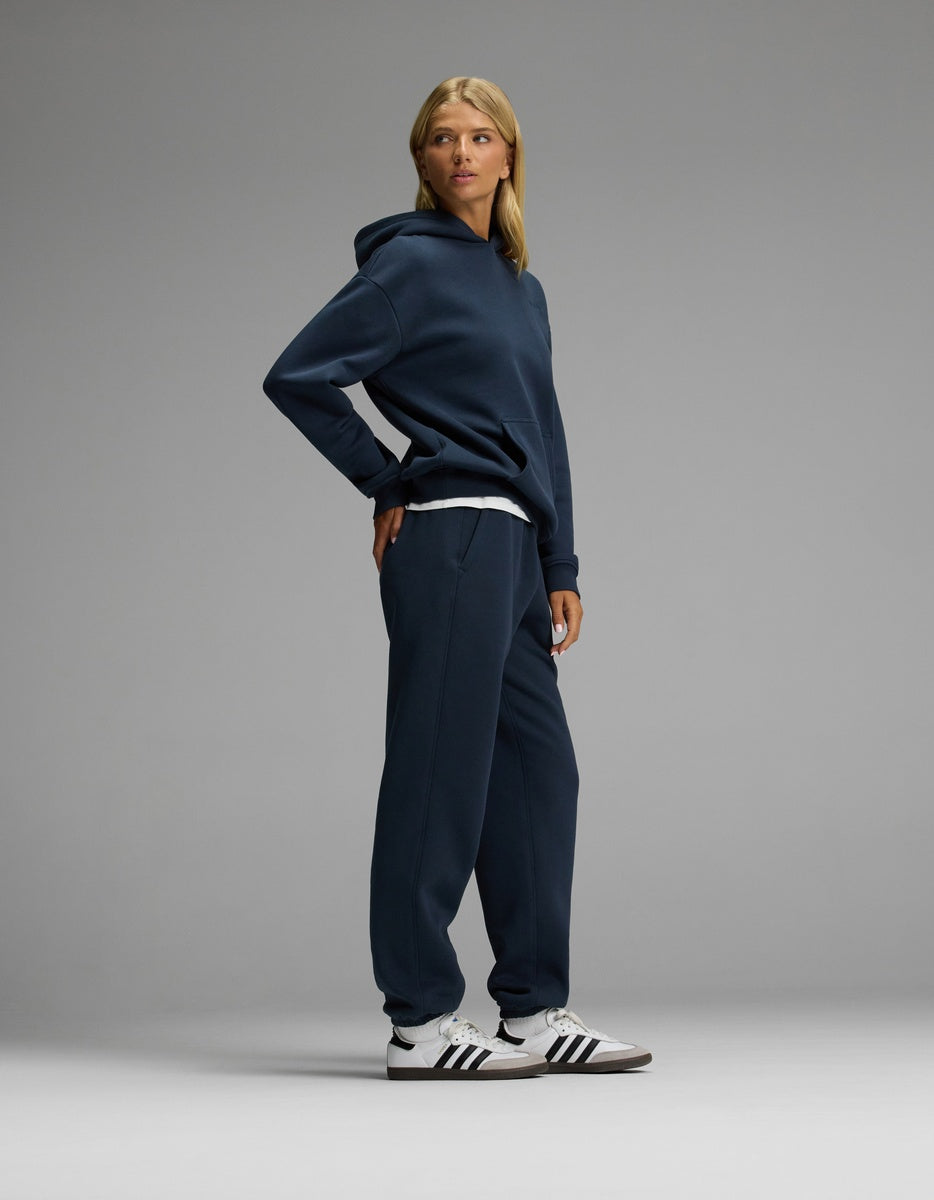 A person stands in a relaxed pose wearing the Cozy Earth Women’s CityScape Hoodie & Sweatpant Set in navy, paired with white Adidas sneakers featuring black stripes, against a plain gray background.