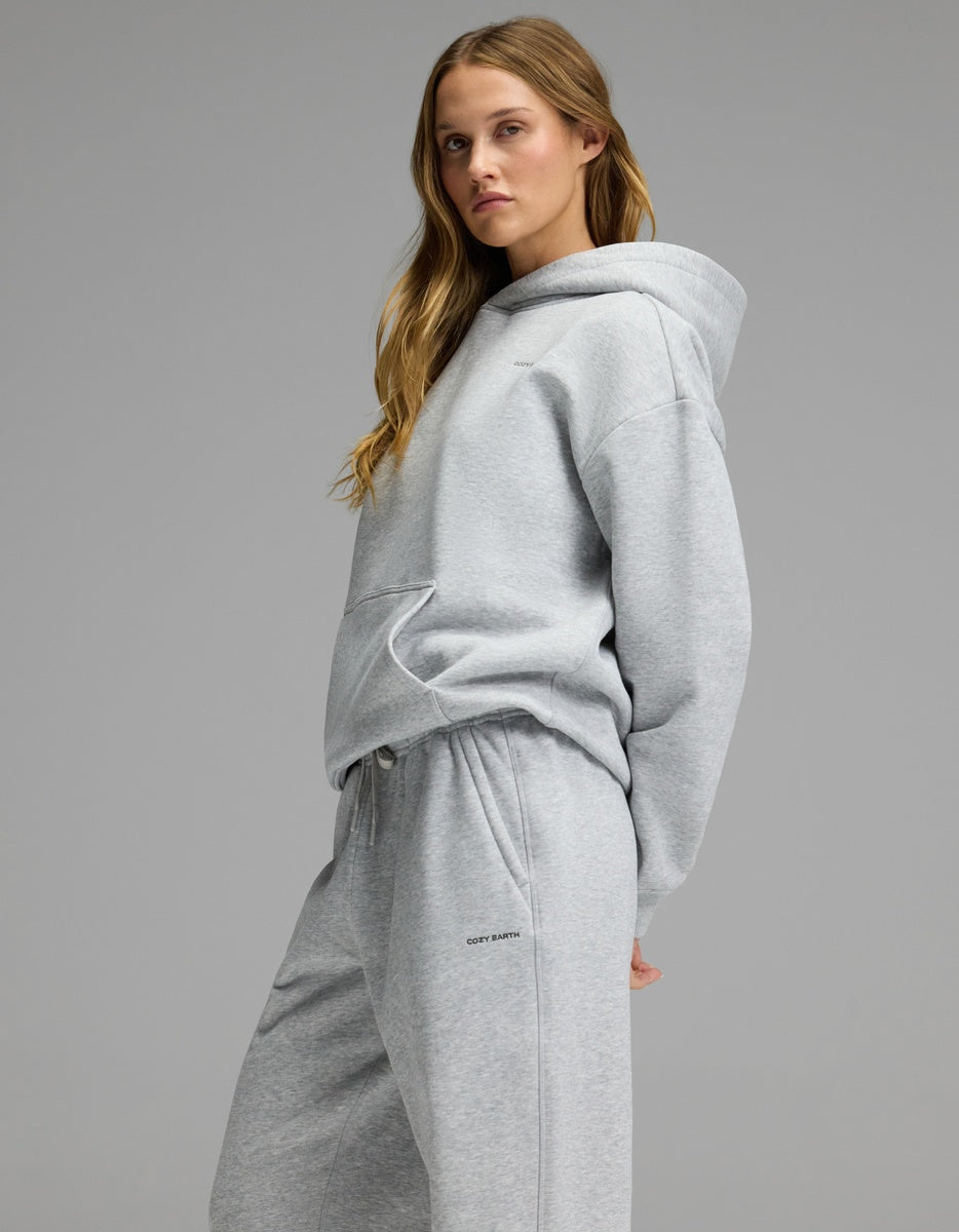 A woman models the Cozy Earth Women’s CityScape Hoodie & Sweatpant Set in light gray, standing with one hand behind her back and gazing sideways against a plain gray backdrop.