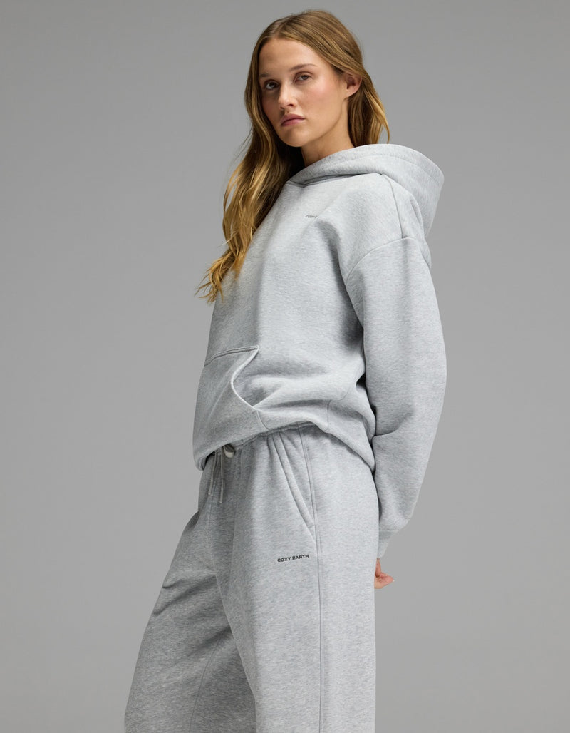 A woman models the Cozy Earth Women’s CityScape Hoodie & Sweatpant Set in light gray, standing with one hand behind her back and gazing sideways against a plain gray backdrop.
