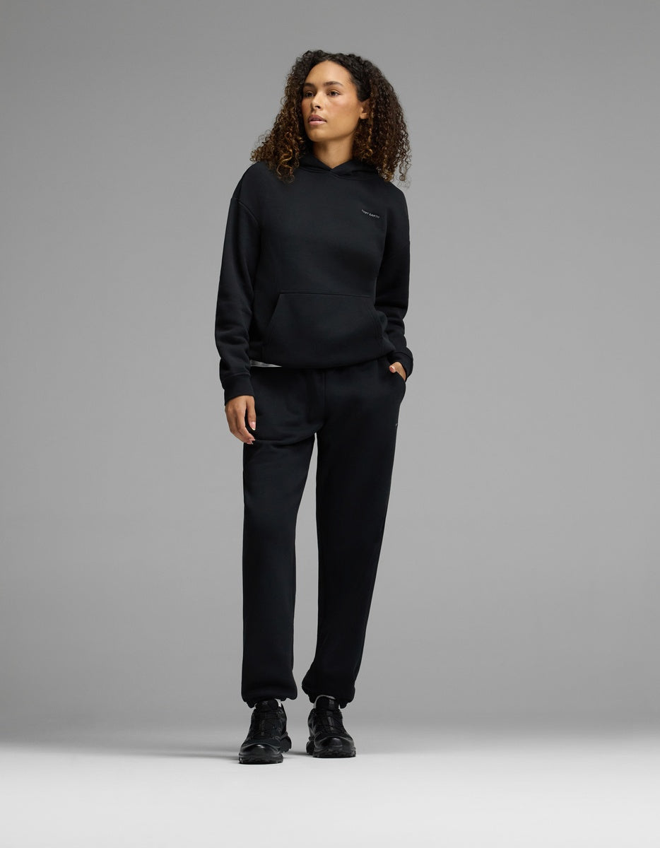 A person with curly hair stands against a gray background, wearing the Cozy Earth Women’s CityScape Hoodie & Sweatpant Set in black, paired with black sneakers and one hand in their pocket.