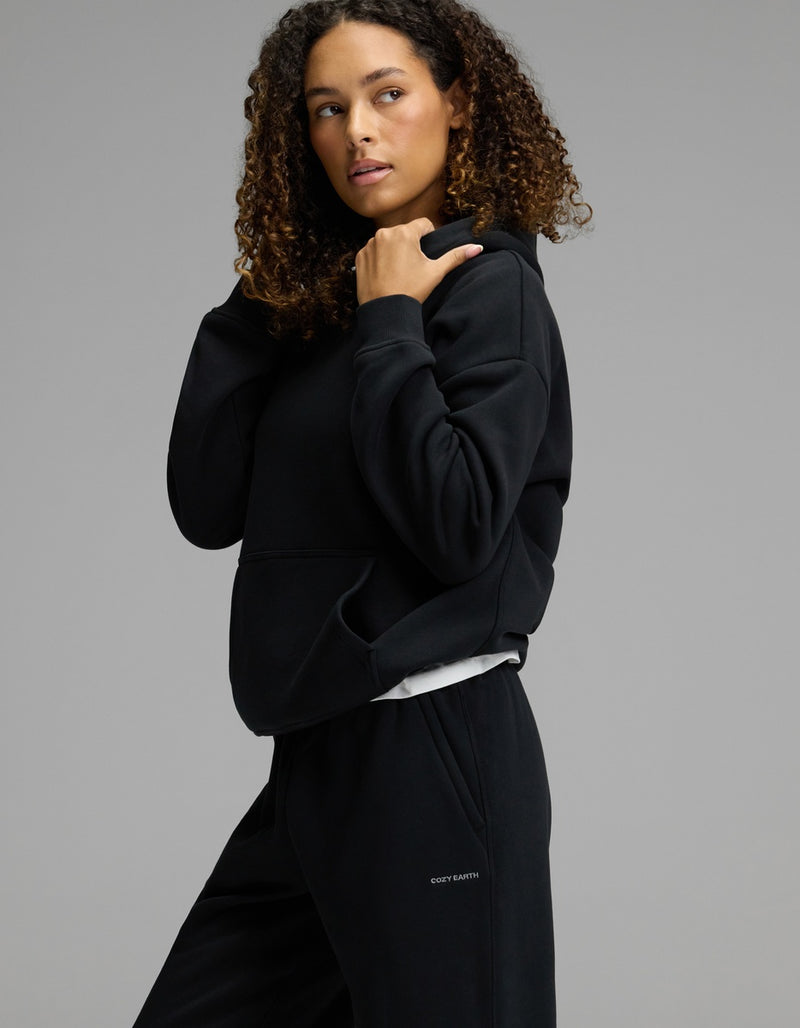 A woman with curly hair wears the Cozy Earth Women’s CityScape Hoodie & Sweatpant Set in black, standing against a neutral gray background and looking to the side as she adjusts her hood.