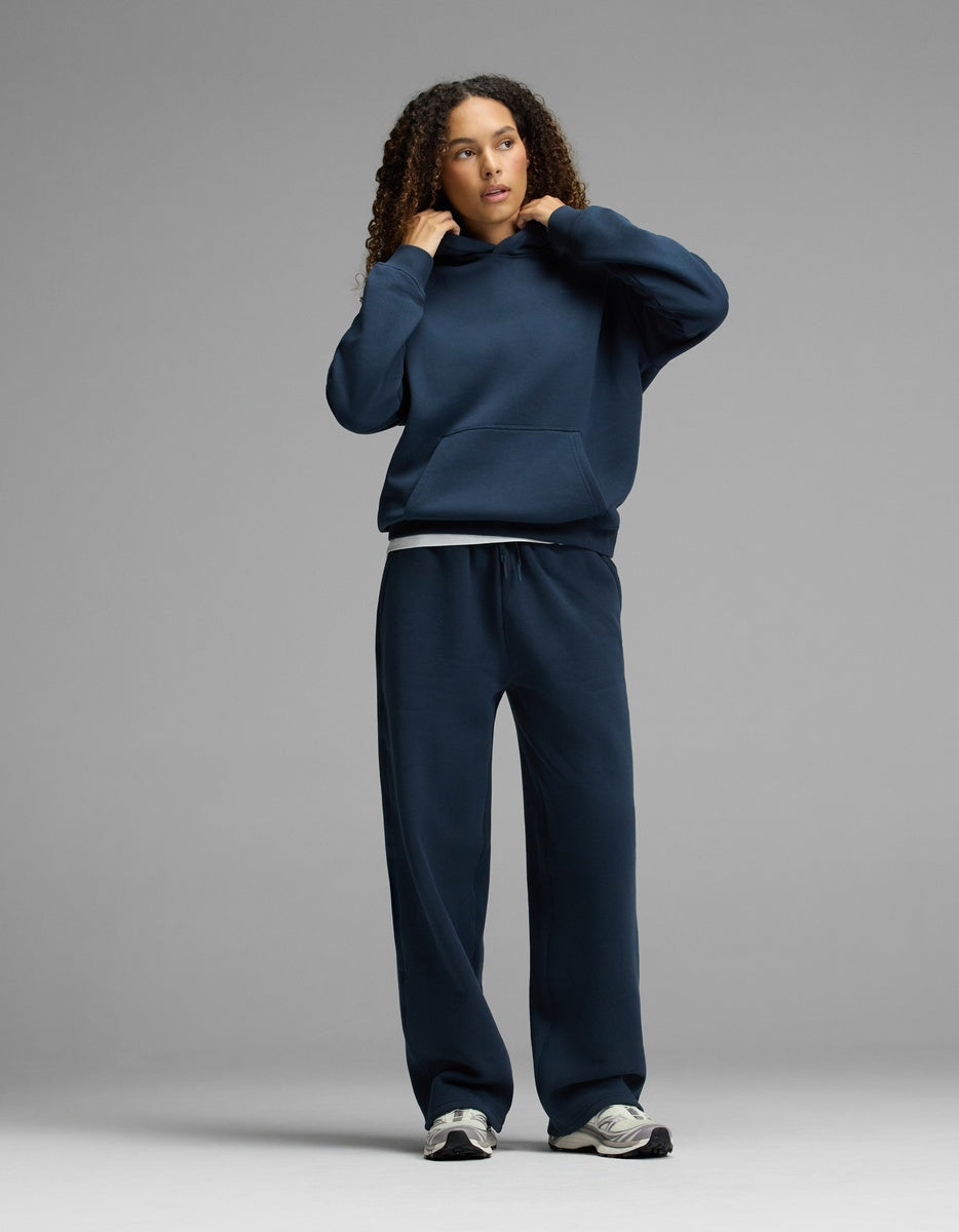 A person with curly hair adjusts the hood of the Women’s CityScape Hoodie & Wide Leg Pant Set by Cozy Earth in navy blue, standing against a plain gray background.