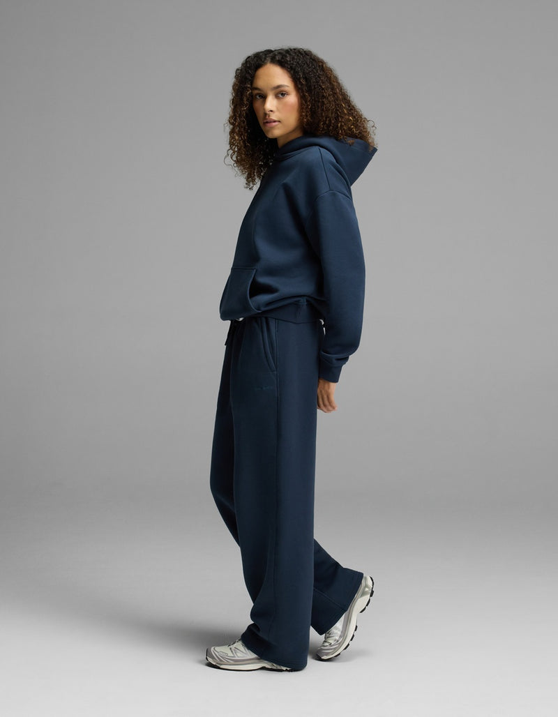 A person with curly hair models the Women’s CityScape Hoodie & Wide Leg Pant Set by Cozy Earth in navy blue, paired with white sneakers and standing sideways against a plain gray background.