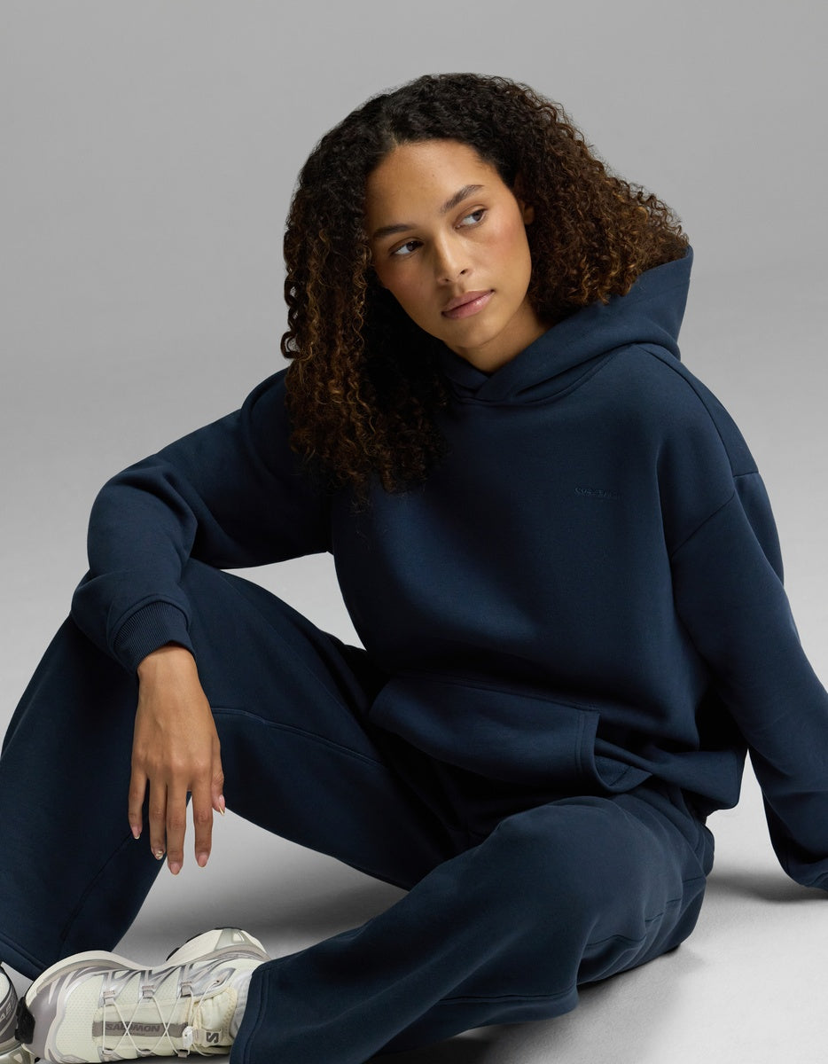 A person with curly hair wears the Women’s CityScape Hoodie & Wide Leg Pant Set by Cozy Earth in navy blue, paired with white sneakers, sitting on a light gray background.