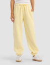 A person stands against a plain background with hands in the pockets of Cozy Earth Women's Cityscape 2.0 Sweatpant in light yellow, paired with a white top and white sneakers.|Color:Meringue