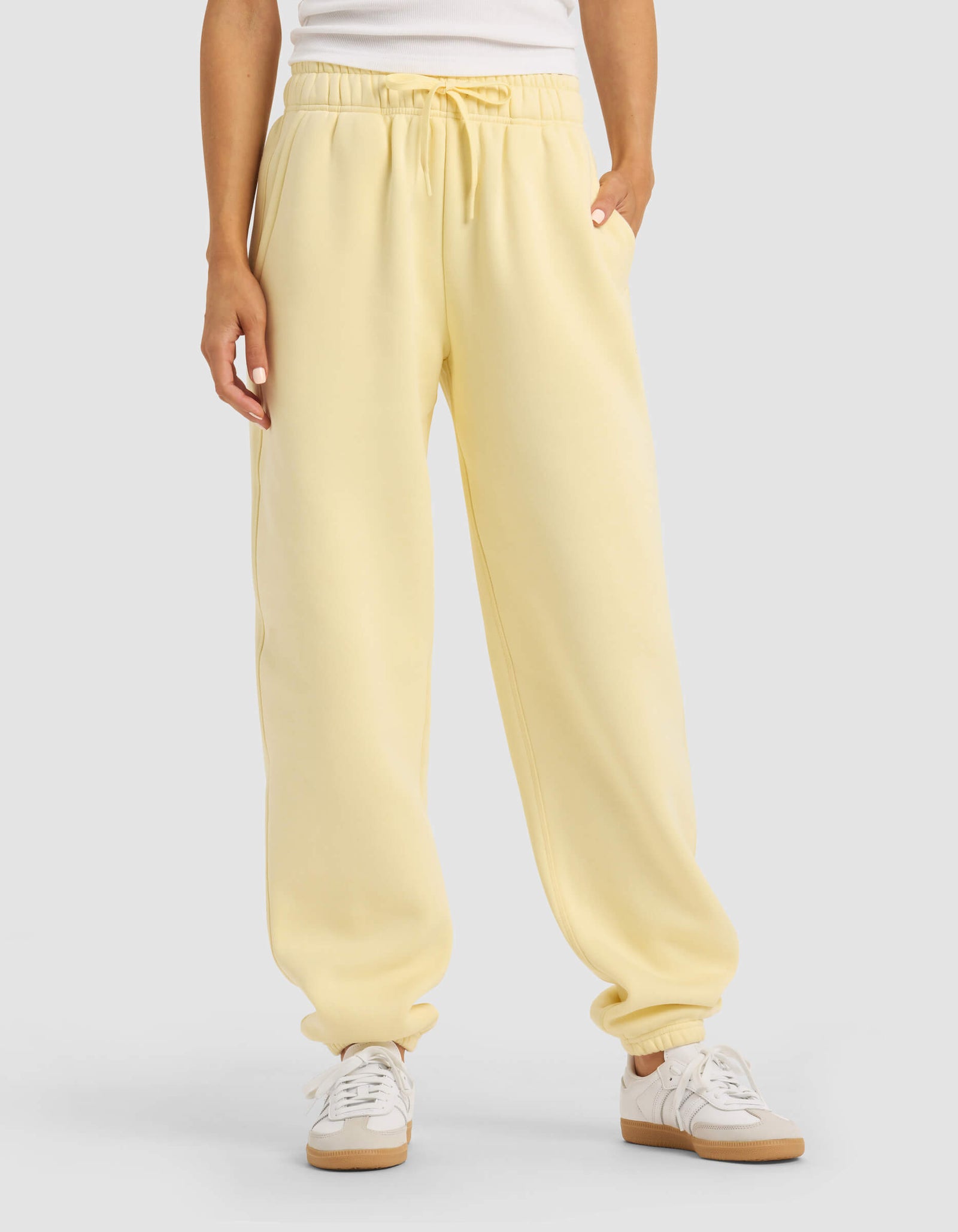 A person stands against a plain background with hands in the pockets of Cozy Earth Women's Cityscape 2.0 Sweatpant in light yellow, paired with a white top and white sneakers.