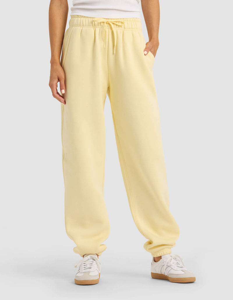 A person stands against a plain background with hands in the pockets of Cozy Earth Women's Cityscape 2.0 Sweatpant in light yellow, paired with a white top and white sneakers.