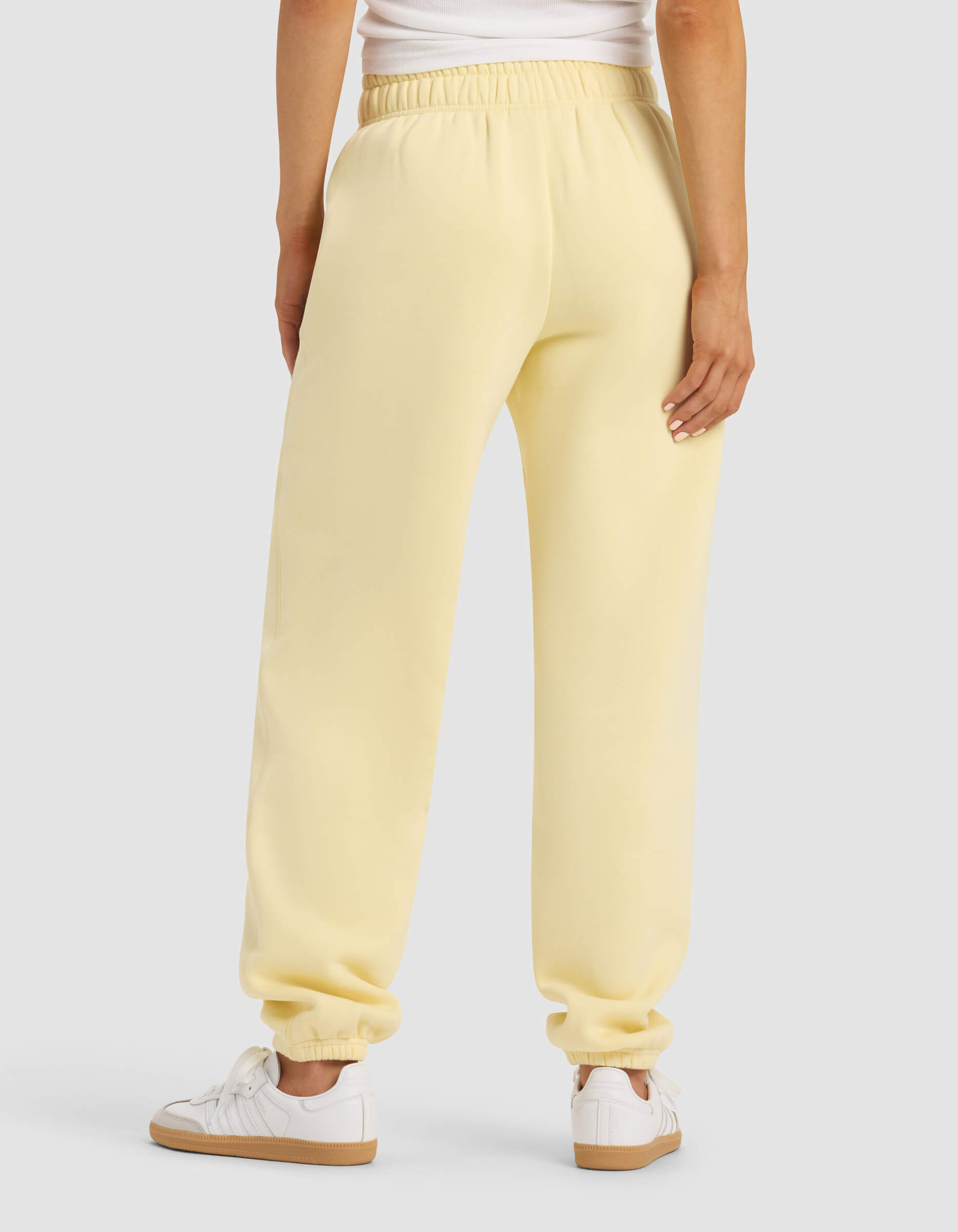 A woman is shown from the back wearing Cozy Earth’s Women's Cityscape 2.0 Sweatpant in light yellow, paired with a white top and white sneakers, standing against a plain light gray background.