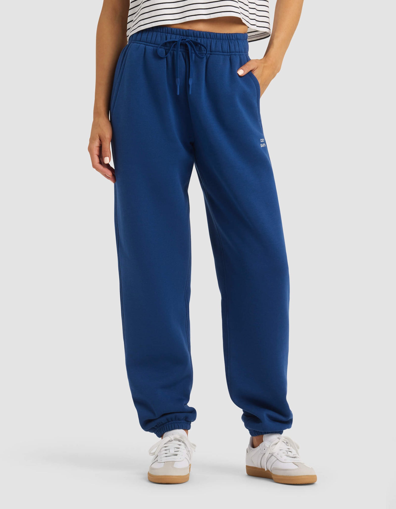 A person wearing Cozy Earth's Women's Cityscape 2.0 Sweatpant in royal blue, paired with a striped crop top and white sneakers, stands against a plain background, showing only their lower torso and legs.