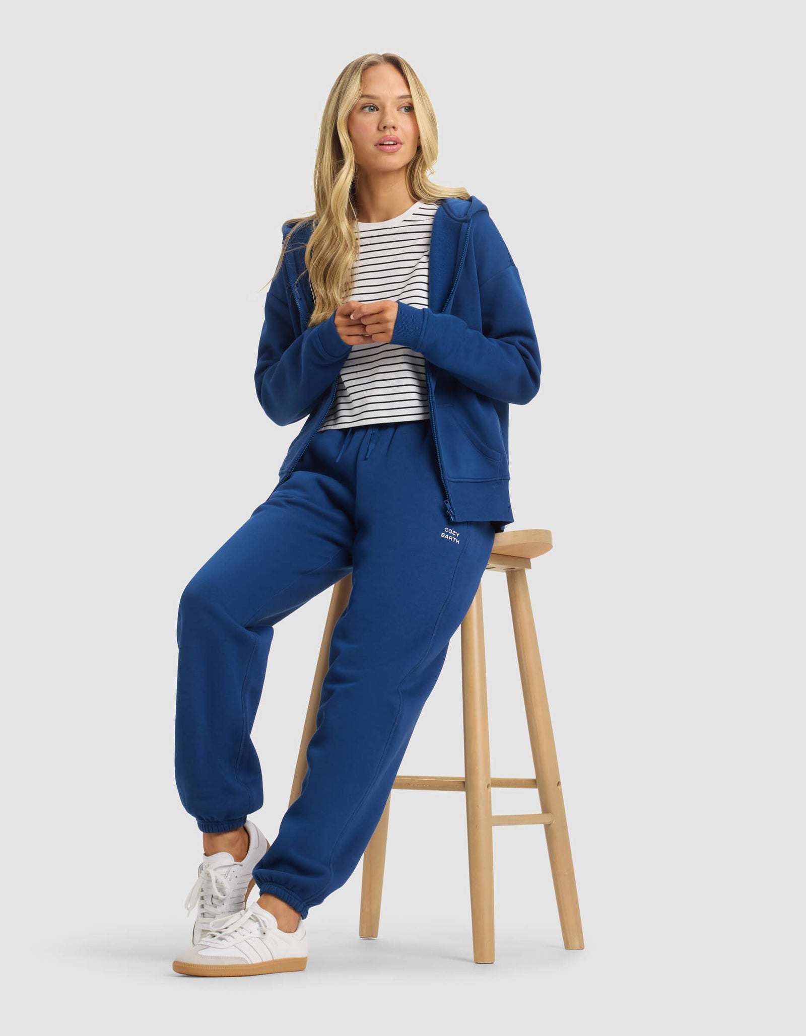 A woman with long blonde hair wears Cozy Earth's Women's Cityscape 2.0 Sweatpant in blue, paired with a white and black striped shirt and white sneakers. She sits on a wooden stool, hands together, looking slightly to the side.